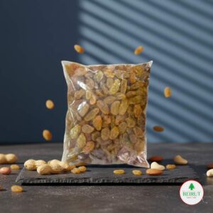 Bolsa de plástico transparente llena de uvas pasas rubias sobre una tabla de pizarra negra, con algunas pasas y frutos secos alrededor.