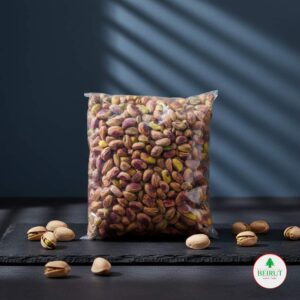 Bolsa de plástico sellada con pistachos naturales, color verde y morado, sobre una superficie oscura de pizarra con algunos pistachos en cáscara dispersos.