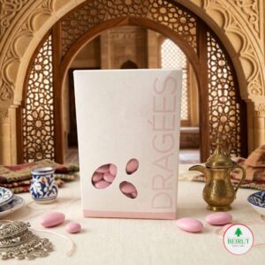 Caja blanca de Dragées con diseño minimalista y ventana que muestra almendras confitadas rosadas, dispuesta en una mesa con decoración árabe tradicional y una cafetera de bronce al fondo.