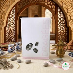 Caja blanca de Dragées con diseño minimalista y ventana que muestra almendras confitadas grises, dispuesta en una mesa con decoración árabe tradicional y una cafetera de bronce al fondo