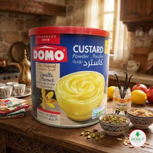 Una lata azul de natilla en polvo marca DOMO (Custard Powder) con sabor a vainilla, situada sobre una mesa de madera rústica. La lata muestra una imagen de un bol con natilla amarilla cremosa.