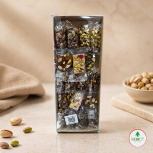 Caja vertical transparente con surtido de dulces árabes de pistacho individuales sobre una mesa con pistachos sueltos.